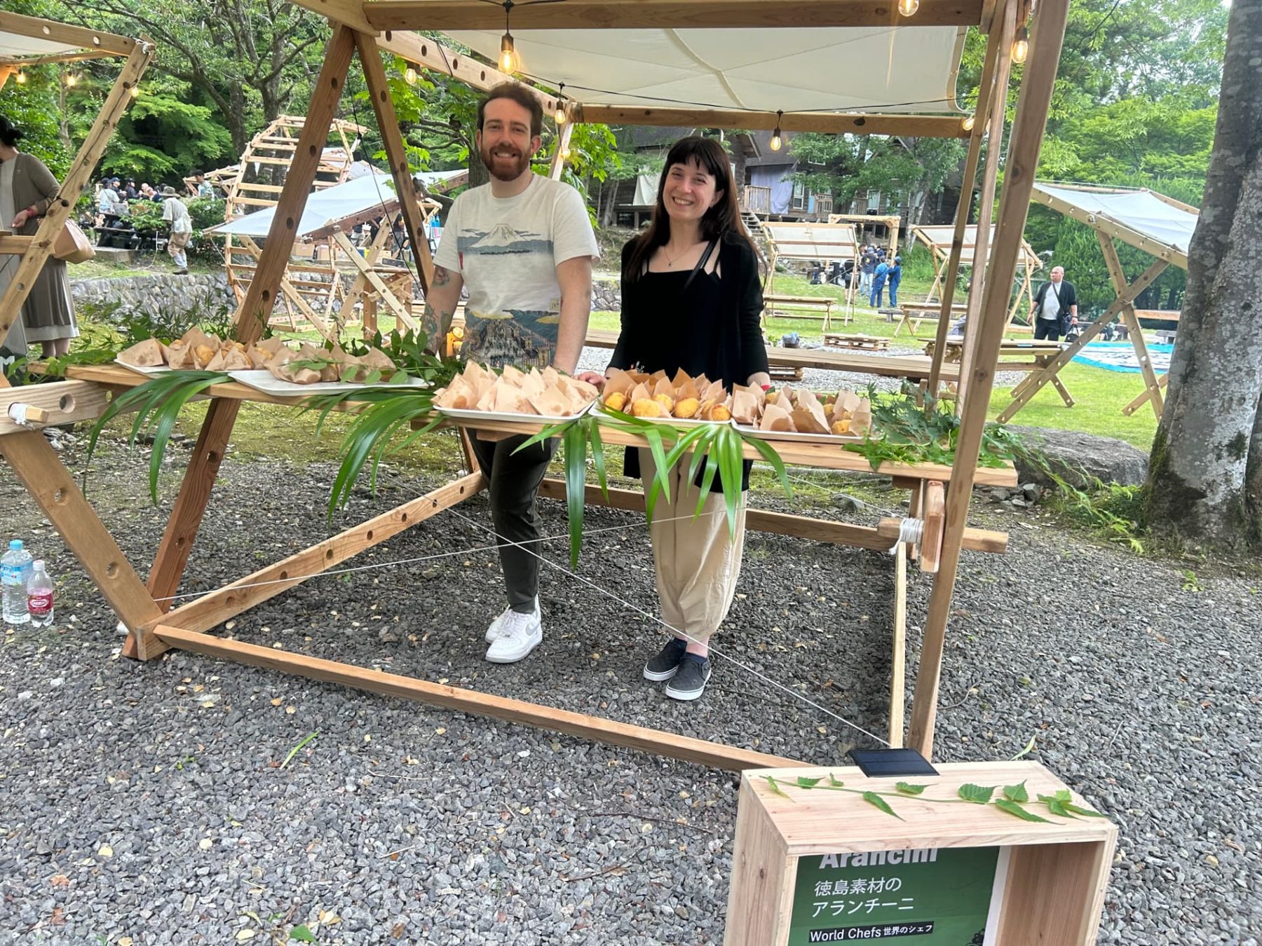 A chef and colleague behind a wooden food stall displaying arancini by day