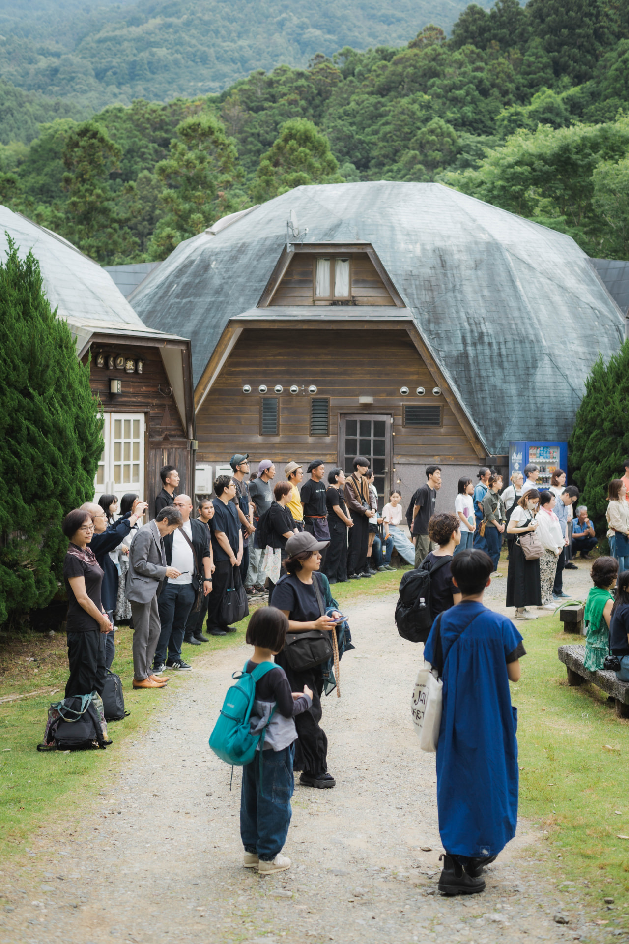 A crowd gathered on the path between the dome-shaped wooden halls and the forested hill