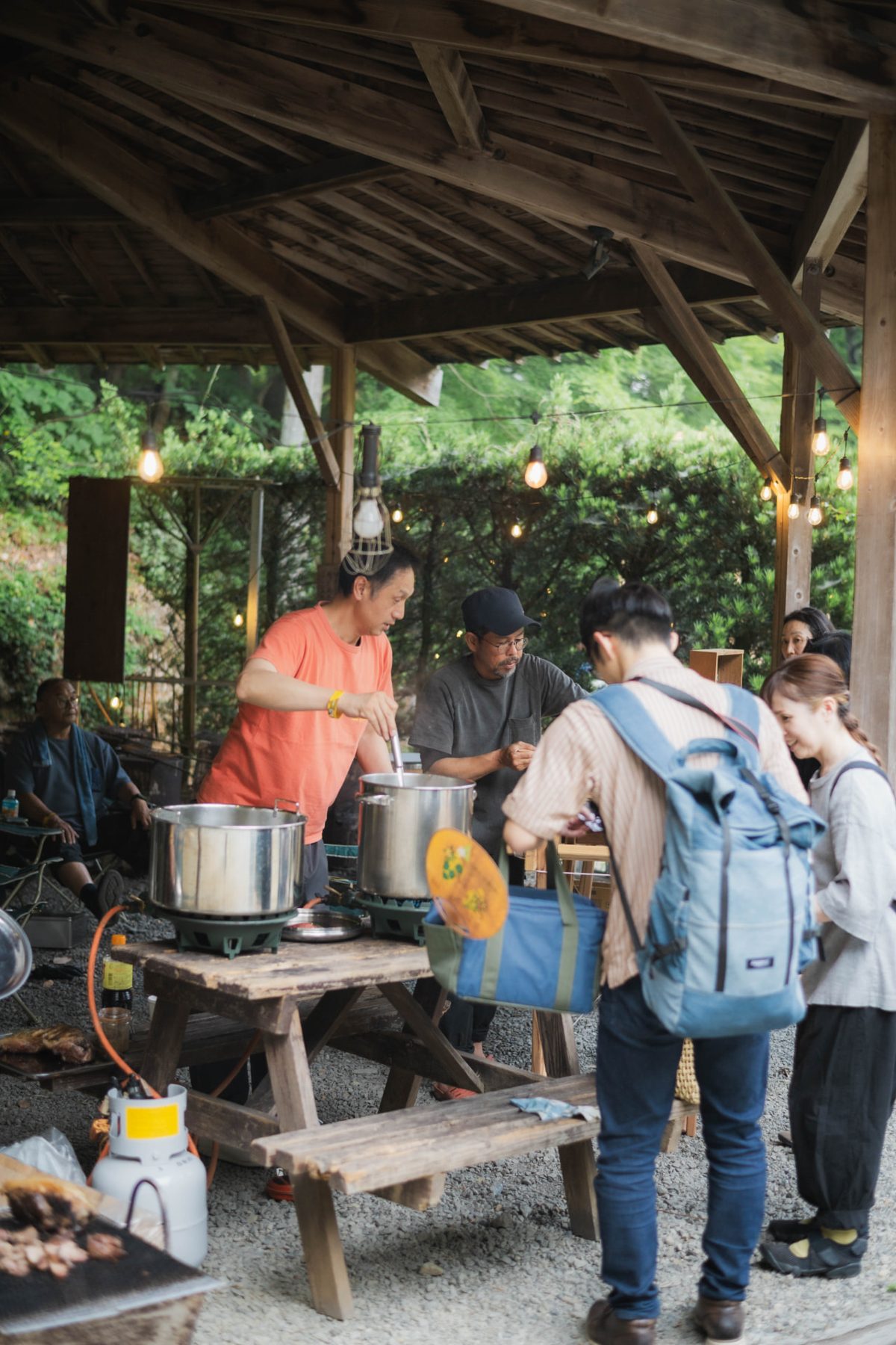 Cooks working over large stockpots under a wooden pavilion in the woods