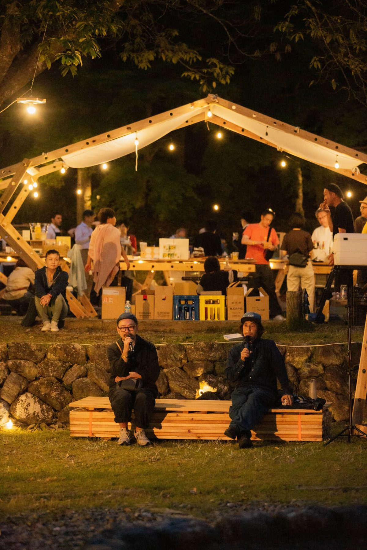 Speakers on a wooden bench at a nighttime gathering under an illuminated canopy
